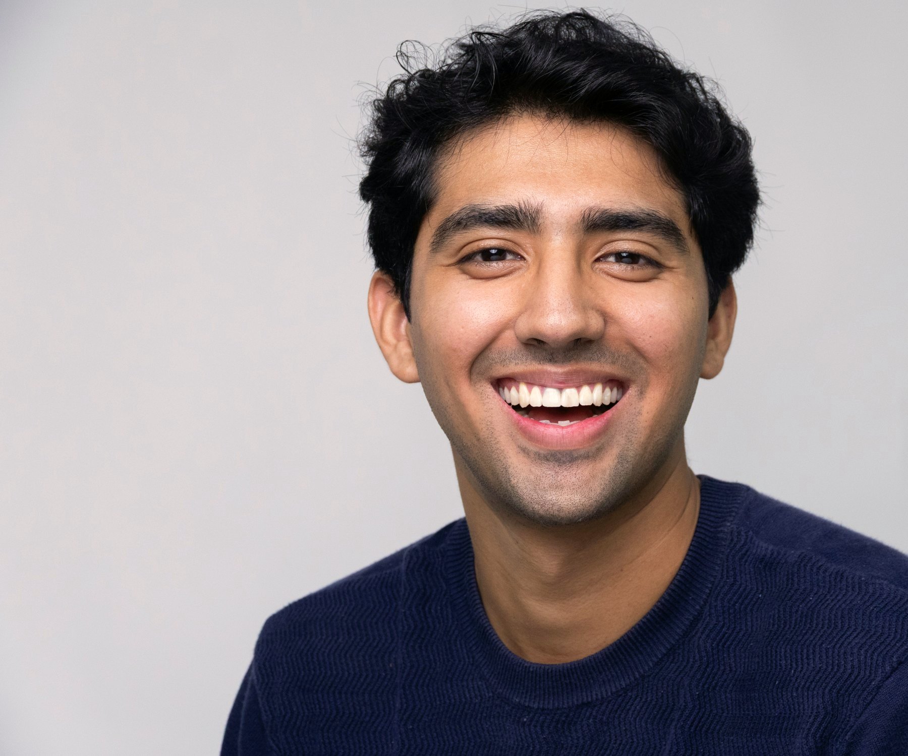 Young man with dark wavy hair laughing, wearing a navy sweater against a soft grey backdrop