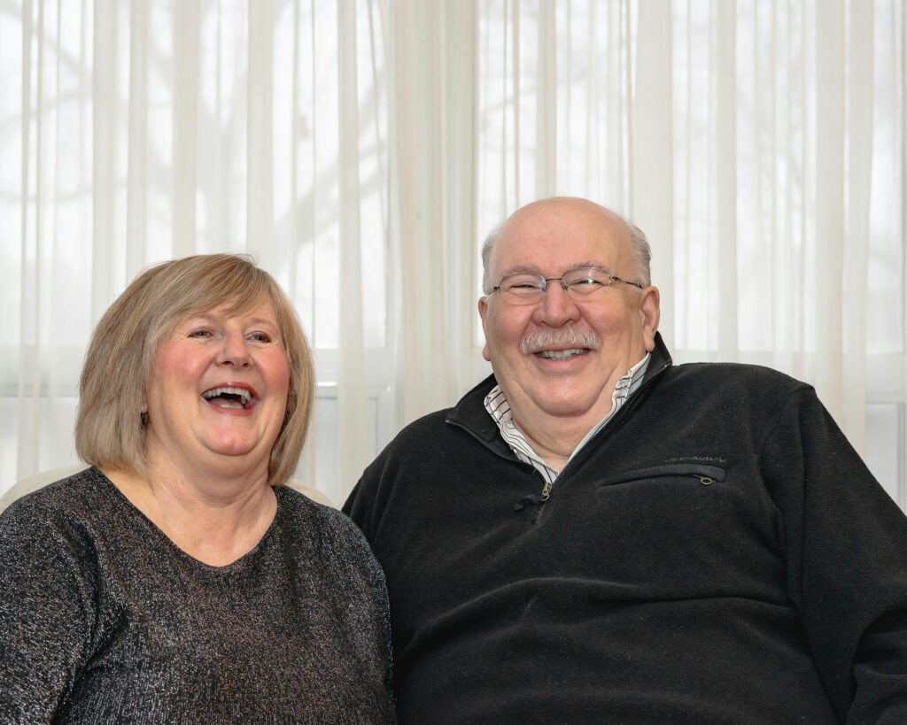 Senior couple laughing together, teeth visible, in a bright living room